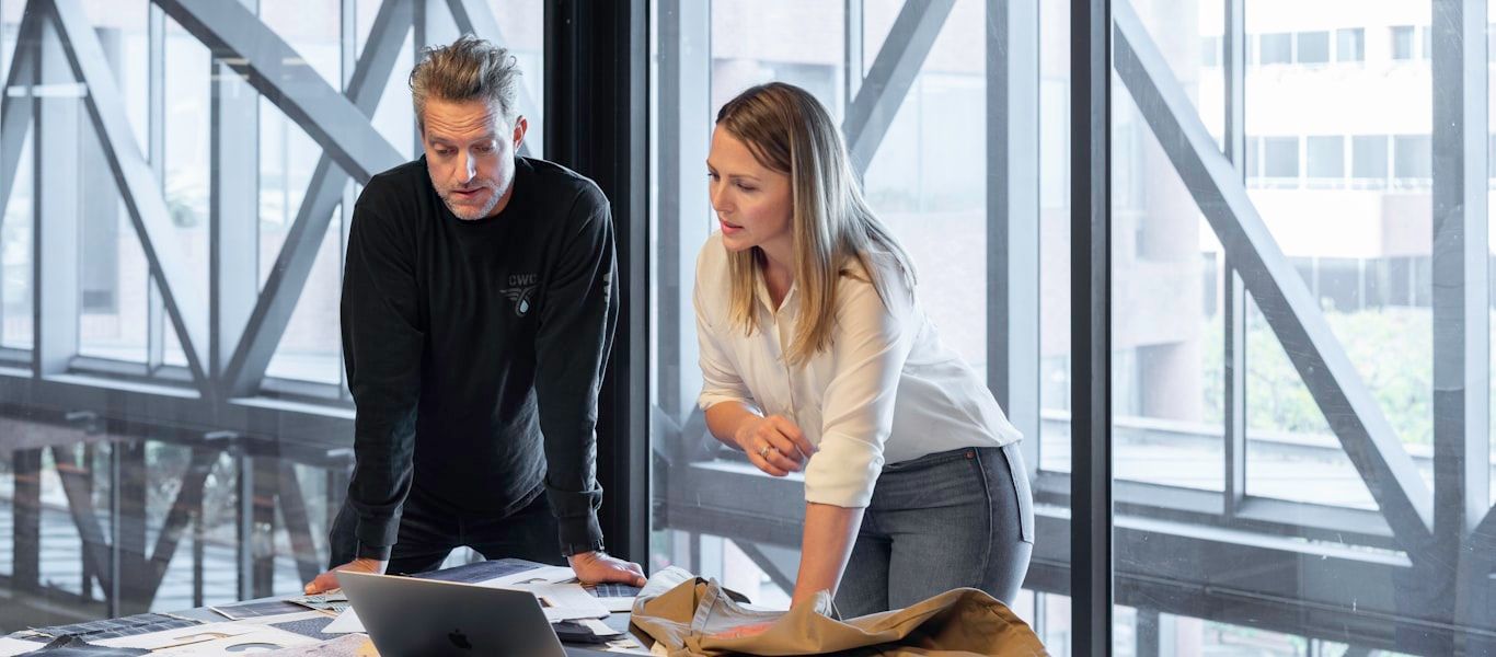 Two colleagues reviewing documents and a laptop together beside an office window